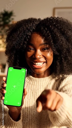 Young woman holding green screen phone in living room with background blur and pointing finger up