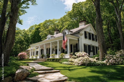 Colonial-style home exterior with a porch, national flag, manicured garden, and forest beyond