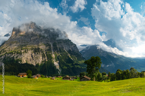 Majestic mountains rise in the background as quaint wooden cabins dot green landscape. A winding path guides a visitor through this serene Swiss alps. Grindelwald Jungfrau Bernese alps Switzerland.