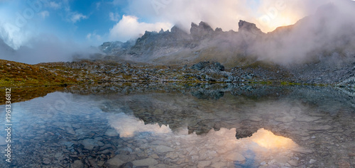 Mist rises over a serene Wildsee lake in the Pizol mountains at sunrise, casting a mystical atmosphere. The rocky terrain and clear waters reflect the early morning light