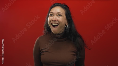 Woman with open mouth gasp and surprised gaze in studio on vivid red backdrop wearing brown turtleneck and hoop earrings; surprise joy energy.