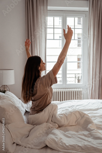 Young woman stretching in bed near window in natural morning light. Slow morning lifestyle.