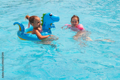 Two young girls enjoying a sunny day in a swimming pool, one on a blue dinosaur float and the other swimming nearby with pink arm floats