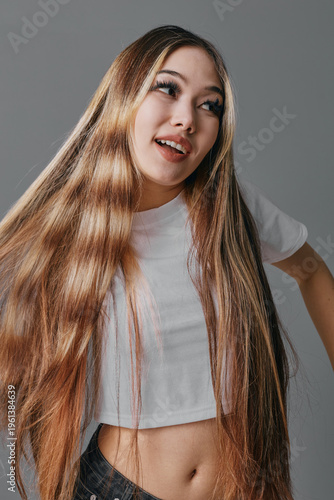 Young woman with long straight hair wearing white crop top and jeans posing with smile on gray background. Casual fashion, beauty, lifestyle, studio portrait with natural light.