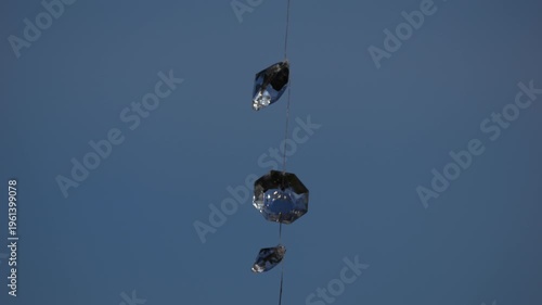 4K telephoto-style shot of decorative glass crystals suspended on a string, gently swaying in the wind against a clean blue sky background. The composition emphasises minimalism, negative space, and s