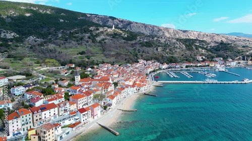 Aerial view of Baska, Krk Island, Croatia. Traditional red-roofed homes hug the turquoise Adriatic Sea, backed by rugged mountains. A picturesque early spring scene.
