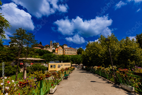 Flower-adorned bridge connects modern path to historic sights in Jajce under bright summer sky