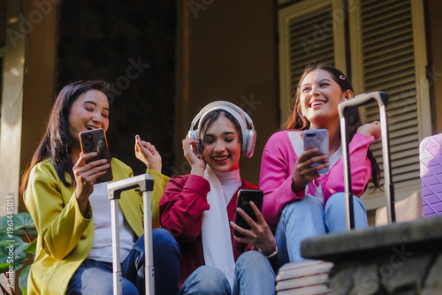 Three delighted young women sit together with their luggage. They are engrossed in their smartphones, listening to music, and radiating happiness as they plan their adventure.