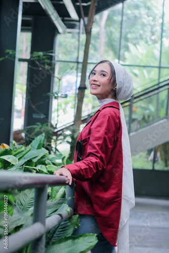 A portrait of a radiant asian woman wearing a white hijab and red jacket, her infectious smile mirroring the thriving nature around her. She stands indoors, her eyes sparkling with happiness.