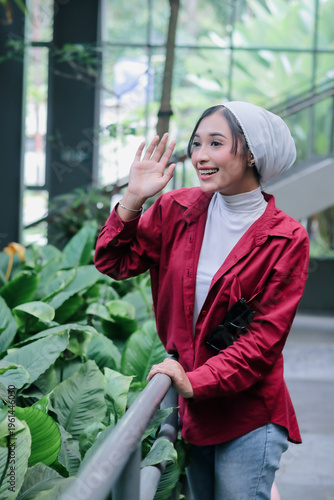 A cheerful woman wearing a hijab smiles as she waves hello in a vibrant setting. Her radiant expression creates a positive atmosphere, inviting the viewer to connect with her.