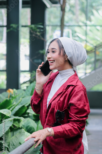 A smiling woman wearing a hijab engages in a phone conversation, showcasing a blend of cultural tradition and modern life. The scene is vibrant and inviting, with a backdrop of lush green foliage.