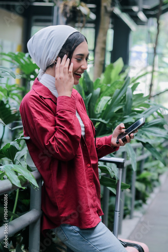 Radiant young businesswoman in hijab exudes happiness as she checks her phone, standing amidst vibrant greenery near her luggage, embodying the spirit of modern, carefree travel.