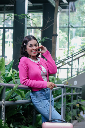 Radiant young woman with a contagious smile, she is ready to begin her travel adventures, posing with her luggage in a vibrant indoor garden. The scene evokes a sense of anticipation and wanderlust.