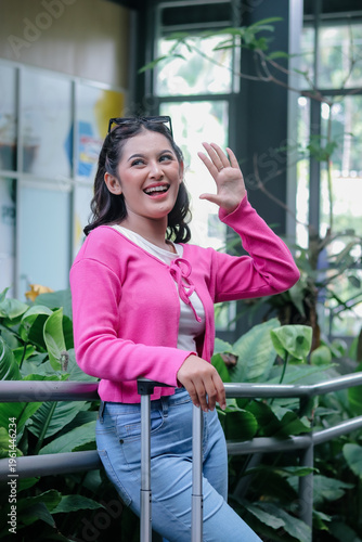 A joyful young woman cheerfully waves goodbye in a modern station, ready for her next journey. She exudes excitement, clutching her luggage, ready for new horizons.