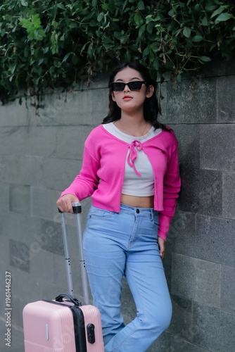 A trendy woman pauses, suitcase beside her, leaning against a textured wall. Dressed in a vivid pink cardigan and light jeans, she exudes effortless urban chic on her anticipated journey.