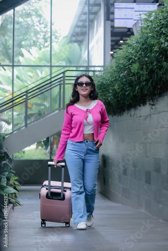 A radiant woman with a pink suitcase embodies modern travel chic, wearing shades and a vibrant pink cardigan. She radiates confidence as she embarks on a stylish urban escapade.