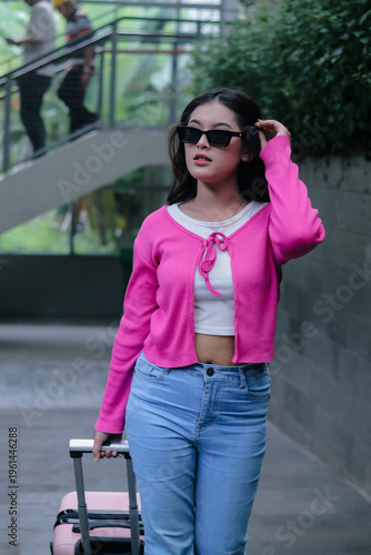 Captured is an elegant young woman with a suitcase, ready for a journey. The casual pink cardigan and trendy sunglasses enhance her confident aura, embodying the spirit of travel.