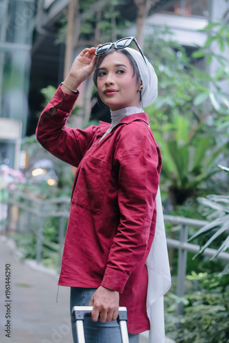 A stylish, confident young woman wearing a hijab poses outdoors with her luggage, ready to embark on a new adventure. She exudes happiness, embracing the promise of her journey.