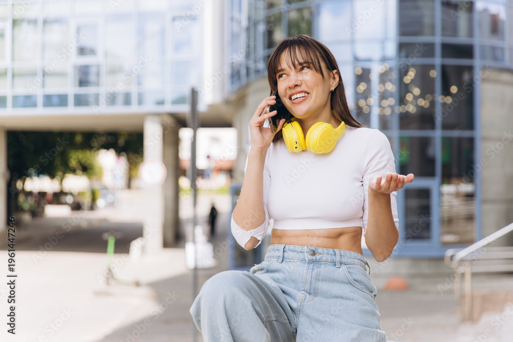 Fototapeta premium Young woman enjoying phone call with casual style, yellow headphones, and smiling expression on city street