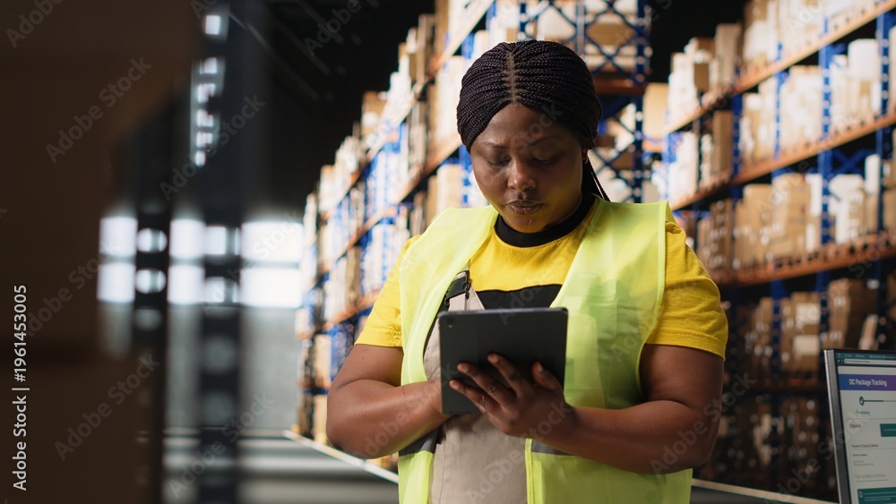 Naklejka premium African american female staff in hi vis vest counting packages on industrial racks, processing custom orders for e-commerce retail operations. Large business management in storehouse. Camera B.
