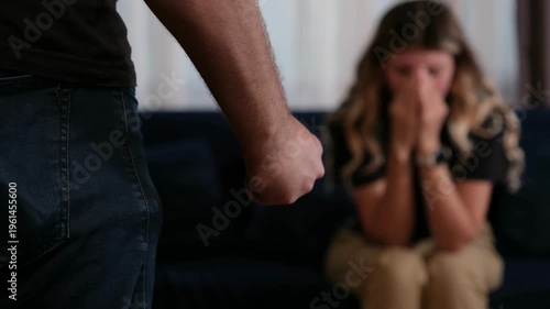 Close up of a man's clenched fist in the foreground with a terrified young woman sitting in the background during a domestic violence or psychological abuse scene indoors at home