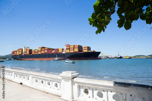 Large container cargo ship departing from Port of Santos, Brazil, with stacked containers, calm sea and coastal landscape on a sunny day.