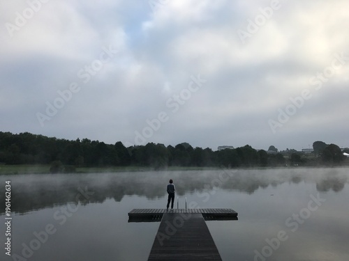 Fotografi Moody and atmospheric lakeside scene featuring a solitary person standing on a wooden pier extending into a calm lake covered with soft mist