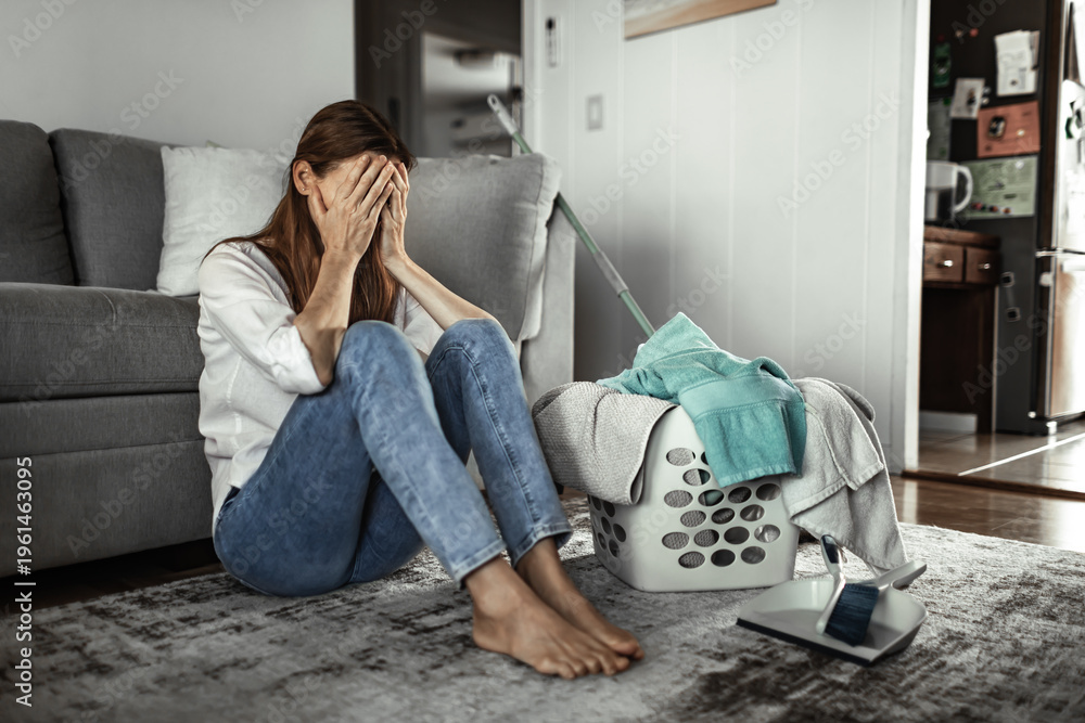 Obraz premium Exhausted woman sitting on floor next to laundry basket feeling overwhelmed by housework, domestic stress and burnout concept 
