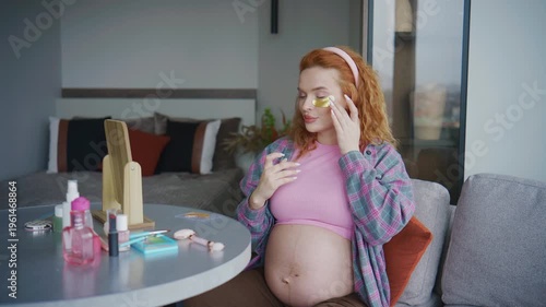 Pregnant Woman with red hair applies makeup while sitting on a sofa in a modern living room with a large window during the daytime