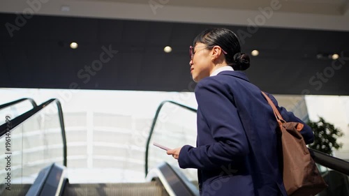 Young Japanese businesswoman riding escalator with phone and bag