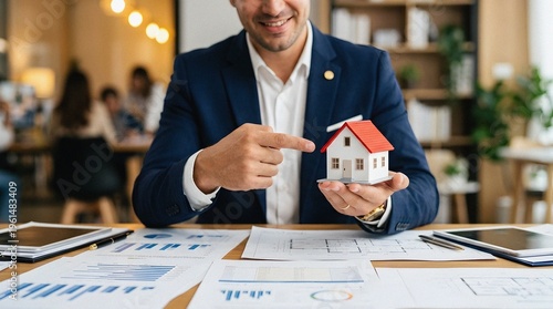 Smiling real estate agent pointing at a house model sitting at a desk with financial documents in a modern office.