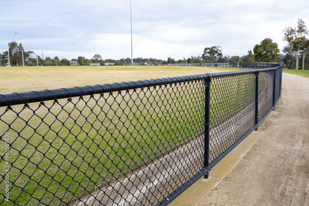 Obraz premium Chain link fence along sports field with grassy oval and walking path in suburban Melbourne Australia. Community sports infrastructure. Enclosed public recreational space for local activity