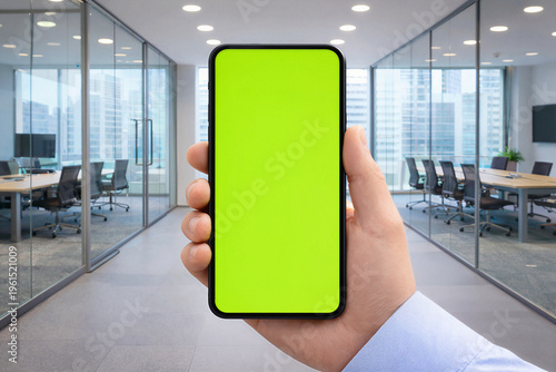Hand holding a smartphone with a green screen in a modern office interior. Perfect mockup for apps, websites, and digital presentations, offering clean space for creative content and branding.