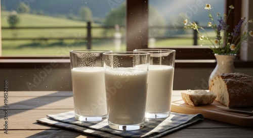 Three tall glasses filled with fresh, white liquid, likely milk, sit on a wooden table adorned with a checkered napkin, accompanied by a loaf of bread and a vase of flowers.