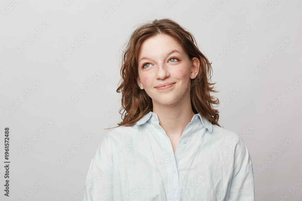 Fototapeta premium Young woman with wavy brown hair wearing white shirt looks up thoughtfully on light background. Concept of contemplation, hope, and casual lifestyle portrait with natural expression.