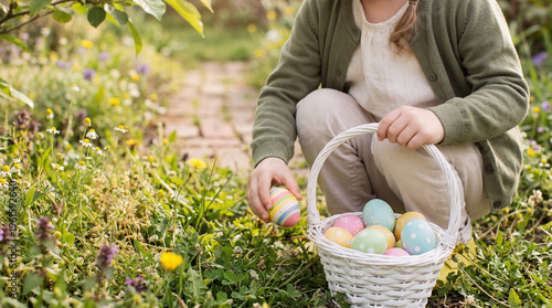 Cute young girl on an exciting outdoor Easter egg hunt, placing a beautifully decorated egg into her white basket.