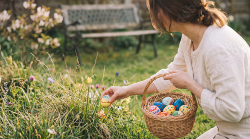 Woman collecting colorful decorated Easter eggs in a wicker basket from lush garden grass on a sunny day.
