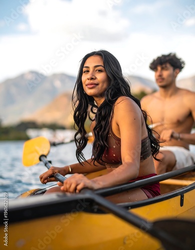 A woman in a swimsuit smiles, kayaking with a man on a scenic lake near mountains