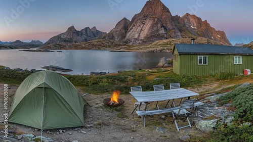 Camping scene at dusk with tent bonfire table and mountains reflected in the water