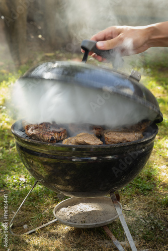 Grilling Meat on a Barbecue in a Backyard on a Sunny Day While a Person Lifts the Lid to Check the Food