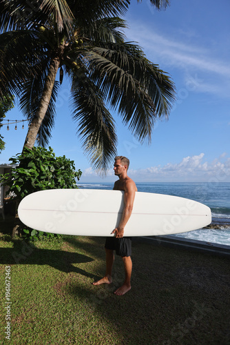 Gentle Paced Surfer Carrying Longboard Walks Past Palm Trees Towards Surf Spot With Serene Blue Sky Overhead