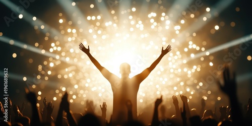 A crowd cheers with raised hands towards a brightly lit stage where a person stands with arms outstretched amid sparkling lights.