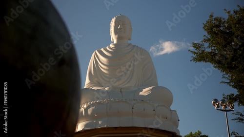 Large white Buddha statue sits in serene park under clear blue sky in mid-afternoon sunlight with trees nearby and people walking around for leisure