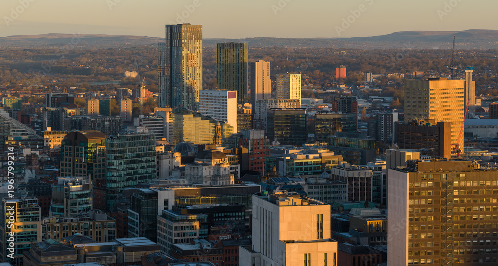 Obraz premium A wide aerial panorama of Manchester city centre at sunrise, featuring modern skyscrapers and office buildings glowing under a golden morning sky.