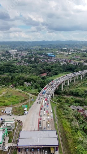 Aerial view of a congested highway curving over a green valley and river, with dense traffic stretching into a small town under a dramatic cloudy sky