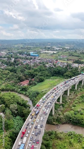An aerial view of a crowded elevated highway crossing a river and lush countryside, with heavy traffic flowing through a town beneath a dramatic cloudy sky