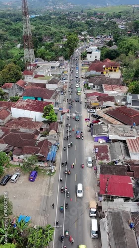 Aerial view of steady traffic on a main road through a small town, with motorcycles and cars passing houses, towers, and lush green surroundings