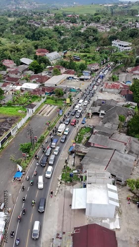 Aerial view of heavy traffic along a winding road through a rural town, with cars and motorcycles passing houses, greenery, and open fields