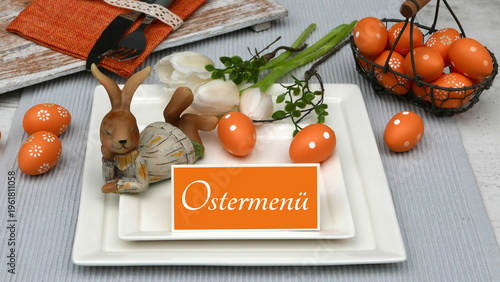 Tischdekoration zu Ostern. Ostertisch mit liegendem Hasen aus Holz, orangefarbenen Ostereiern mit weißen Tulpen und einem Schild mit der Aufschrift 'Ostermenü'