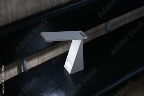 Minimalist white table lamp rests on black surface. Soft glow highlights clean, angular design. Dark background emphasizes modern, sleek aesthetic